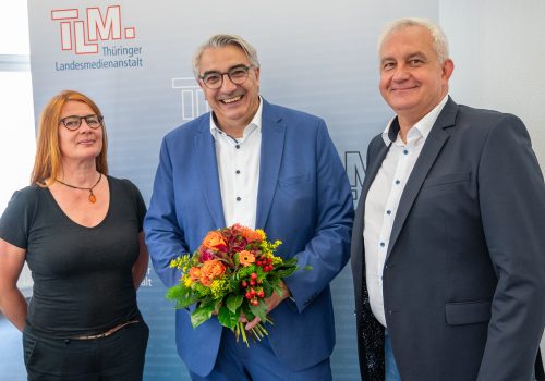 Eine Frau, ein Mann mit Blumenstrauß in der Mitte und ein Mann stehen lächend nebeneinander vor einem TLM-Banner