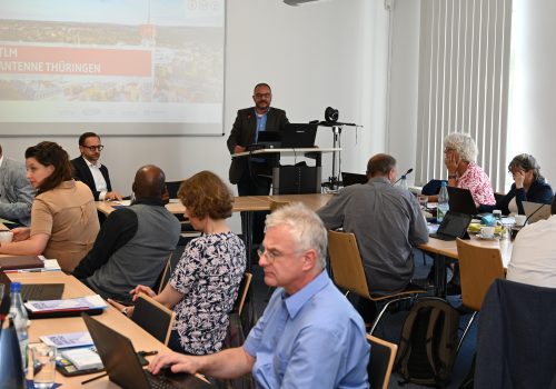 Viele Menschen sitzen an Tischen in einem Sitzungsraum. Ein Mann steht am Rednerpult und redet.