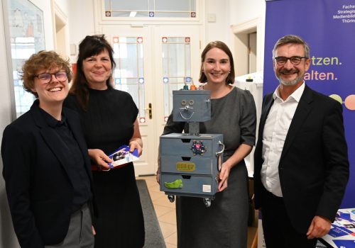 Drei Frauen und ein Mann stehen nebeneinander. Eine Frau hält eine Art Roboter aus Holzkisten in den Händen.