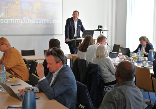 Vielen Menschen sitzen in einem Raum an Tischen und schauen zum Redner.