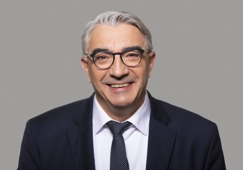 Ein Mann mit weißen kurzen Haaren, weißen Hemd, schwarzen Sakko und schwarzer Brille lächelt in die Kamera.