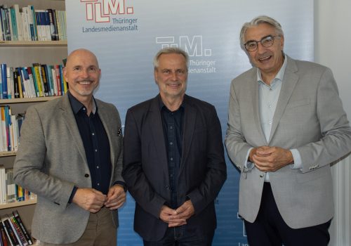 Drei Männer lächeln vor TLM-Banner in die Kamera