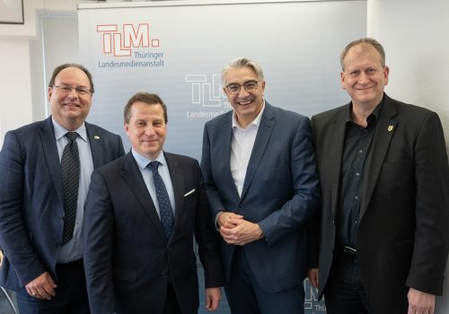 Vier Herren in dunklen Anzügen stehen nebeneinander und lächeln in die Kamera.