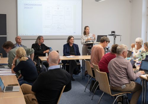 Eine Frau steht am Rednerpult und referiert. Viele Menschen sitzen an Tischen und folgen dem Vortrag.