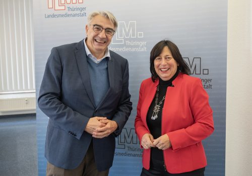 Ein Mann mit blauem Sakko und eine Frau mit rotem Blazer stehen nebeneinander und lächeln in die Kamera.