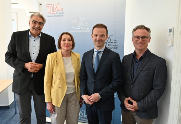 Eine Frau und drei Männer stehen vor einem TLM-Banner und lächeln in die Kamera