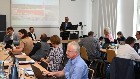Viele Menschen sitzen an Tischen in einem Sitzungsraum. Ein Mann steht am Rednerpult und redet.