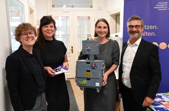Drei Frauen und ein Mann stehen nebeneinander. Eine Frau hält eine Art Roboter aus Holzkisten in den Händen.