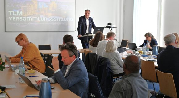 Vielen Menschen sitzen in einem Raum an Tischen und schauen zum Redner.