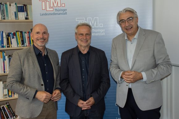 Drei Männer lächeln vor TLM-Banner in die Kamera