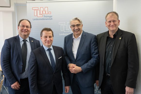 Vier Herren in dunklen Anzügen stehen nebeneinander und lächeln in die Kamera.