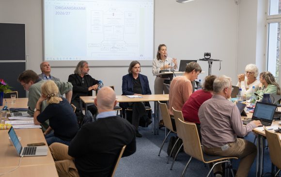 Eine Frau steht am Rednerpult und referiert. Viele Menschen sitzen an Tischen und folgen dem Vortrag.