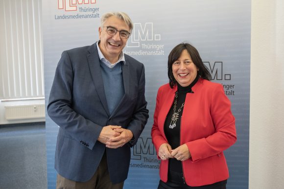 Ein Mann mit blauem Sakko und eine Frau mit rotem Blazer stehen nebeneinander und lächeln in die Kamera.