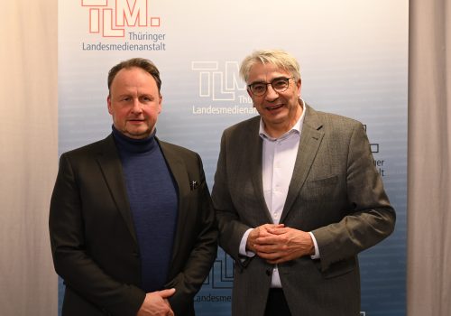 Zwei Männer mit dunklen Sakkos stehen nebeneinander und lächeln in die Kamera.