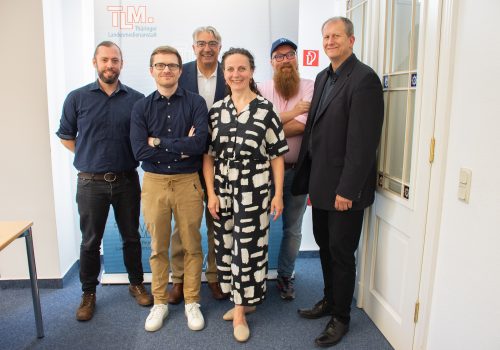 Fünf Männer und eine Frau stehen vor einem TLM-Banner und lächeln in die Kamera