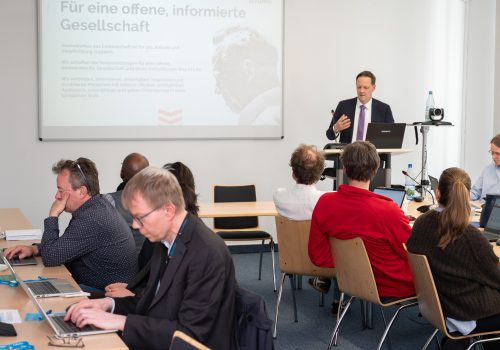 Ein Mann im dunklen Anzug steht an einem Rednerpult vor einer Leinwand und spricht zu Menschen die an Tischen sitzen.