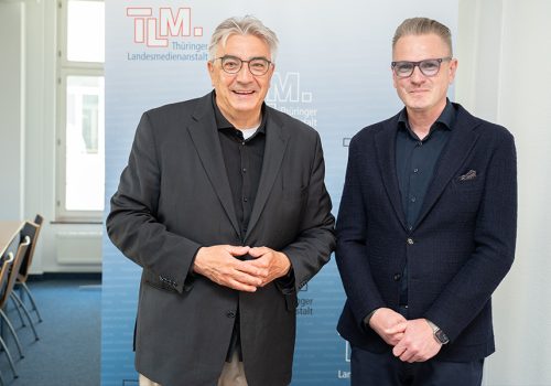 Zwei Männer in dunklen Anzügen stehen nebeneinander und schauen Richtung Kamera.