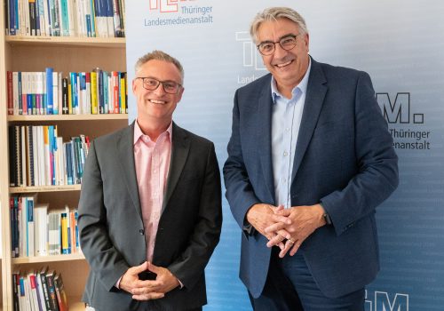 Zwei Männer mit dunklen Anzügen stehen nebeneinander und lächeln in die Kamera.
