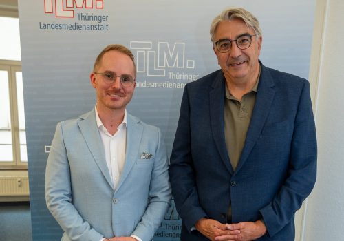 Zwei Männer mit hellblauen und dunkelblauen Sakko stehen nebeneinander und lächeln in die Kamera