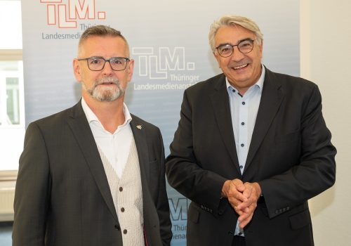 Zwei Männer stehen vor einem TLM-Banner und lächeln in die Kamera