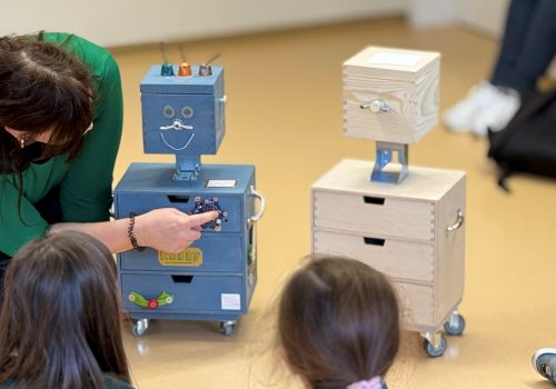 Eine Frau mit dunklem Haar kniet auf dem Boden und zeigt den zuschauenden Kindern die Platine, die am Holzroboter Robby angebracht ist
