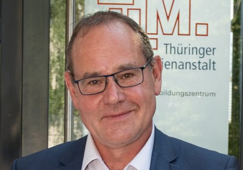 Ein Mann mit weißen Hemd, dunklem Sakko und Brille lächelt in die Kamera.
