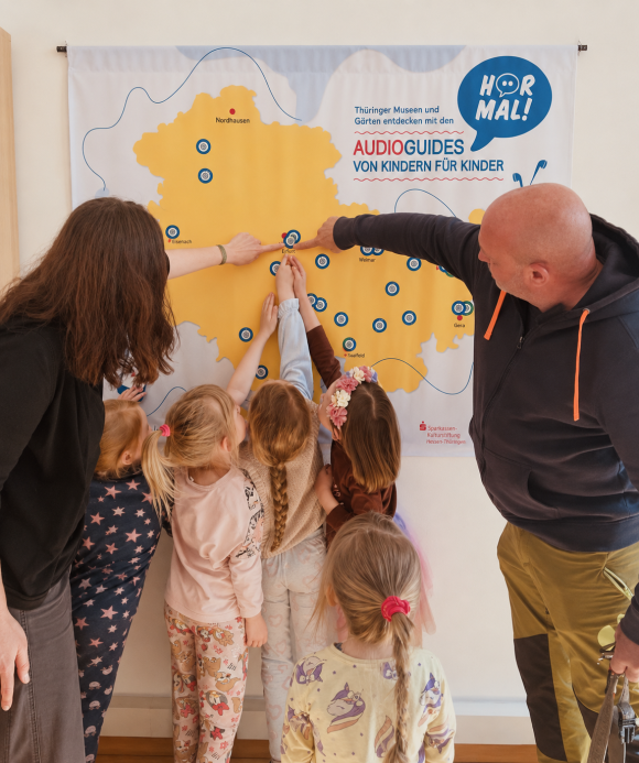 Zwei Erwachsene und fünf Kinder zeigen auf Thüringenkarte mit den Standorten der Hör mal-Projekte