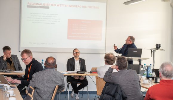 Ein Vortragender steht neben einer Leinwand und spricht mit den Anwesenden.