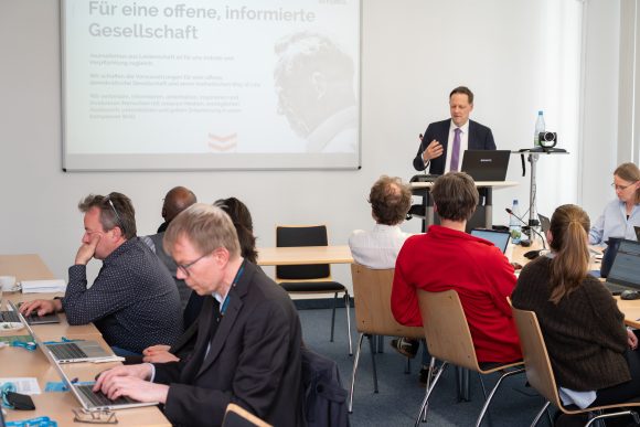 Ein Mann im dunklen Anzug steht an einem Rednerpult vor einer Leinwand und spricht zu Menschen die an Tischen sitzen.