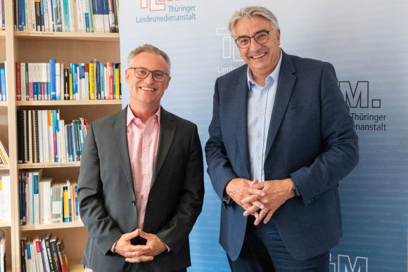Zwei Männer mit dunklen Anzügen stehen nebeneinander und lächeln in die Kamera.