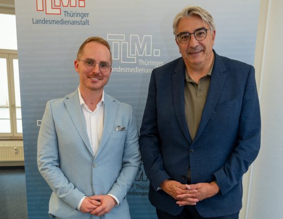 Zwei Männer mit hellblauen und dunkelblauen Sakko stehen nebeneinander und lächeln in die Kamera