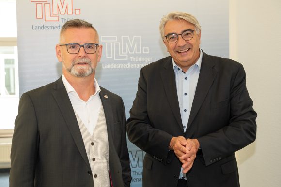 Zwei Männer stehen vor einem TLM-Banner und lächeln in die Kamera