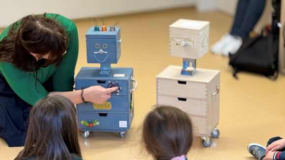 Eine Frau mit dunklem Haar kniet auf dem Boden und zeigt den zuschauenden Kindern die Platine, die am Holzroboter Robby angebracht ist