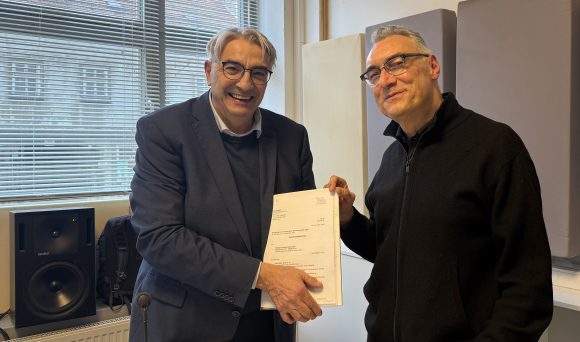 Zwei Männer stehen nebeneinander, lächeln in die Kamera und halten gemeinsam den Fördermittelbescheid im A4-Format fest.