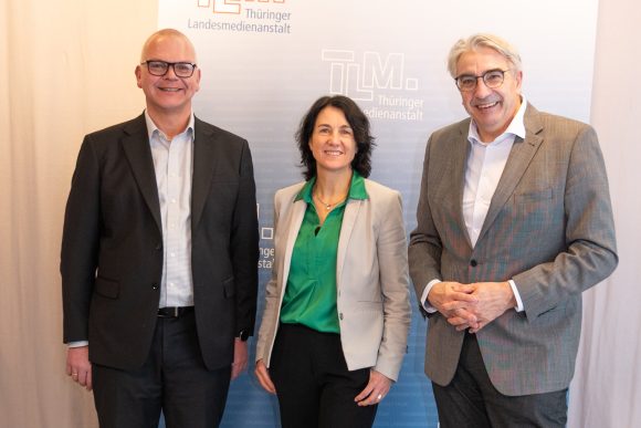 Eine Frau und zwei Männer stehen nebeneinander und lächeln in die Kamera.