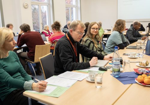 Viele Menschen sitzen in einem Raum an Tischen und beraten sich.