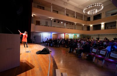 Blick auf Bühne mit Moderatorin und in den Veranstaltungsraum