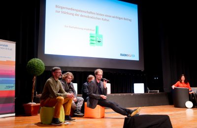 Vier Männer sitzen auf der Bühne, einer spricht ins Mikrofon