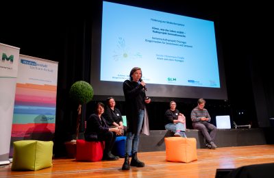 Eine Frau steht mit Mikrofon auf der Bühne ins Mikrofon, vier Personen sitzen dahinter