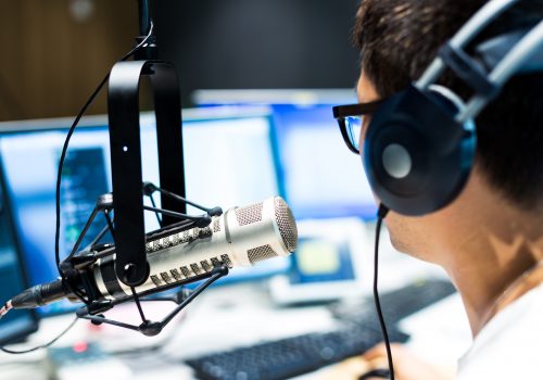 Mann von hinten mit Kopfhörern vor Monitoren am Mikrofon im Radiostudio als JPG